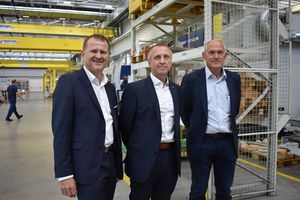 3 men in suits standing in a production hall, smiling for the camera